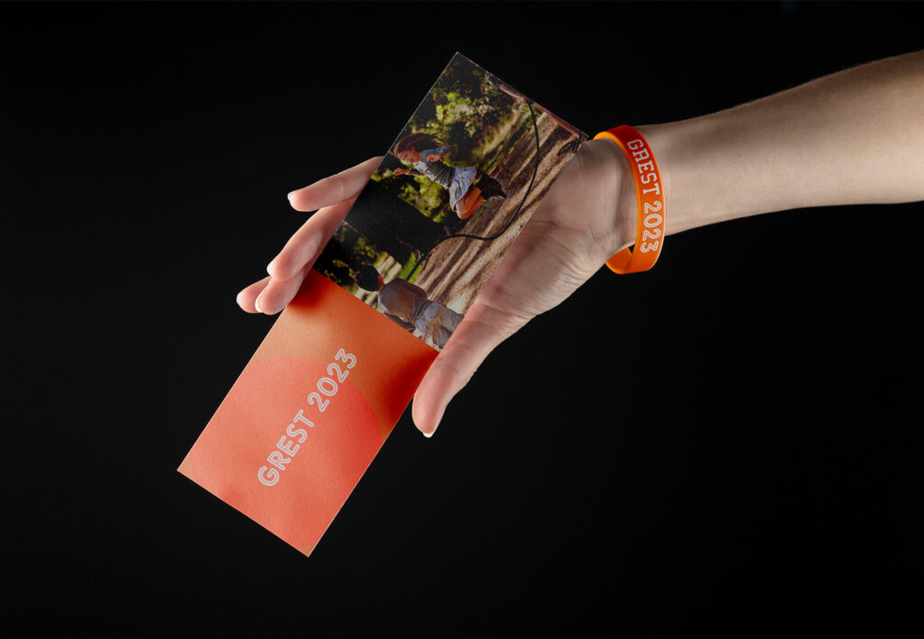 Mano che indossa un bracciale arancione con la scritta Grest2023 e afferra un flyer con scritto Grest2023 e una foto di una bambina che salta la corda