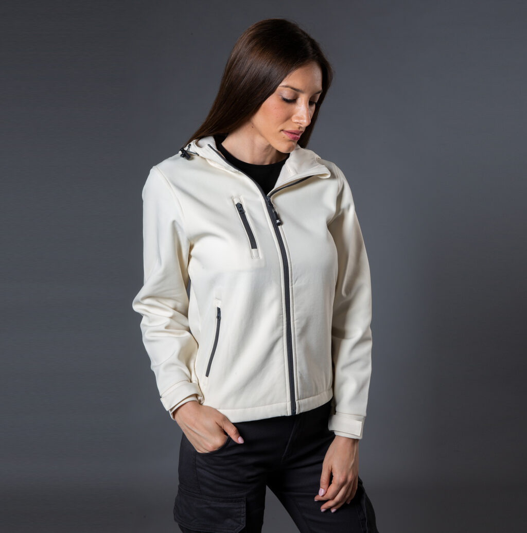 Foto di ragazza castana che indossa uno soft-shell color panna e pantaloni neri. Sfondo studio grigio